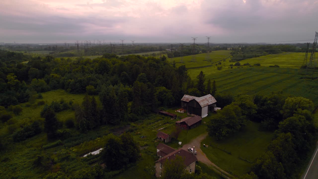 Svelamento cinematografico di una fattoria accanto a una strada di campagna rurale con un campo coltivato e linee elettriche sotto cieli nuvolosi al tramonto