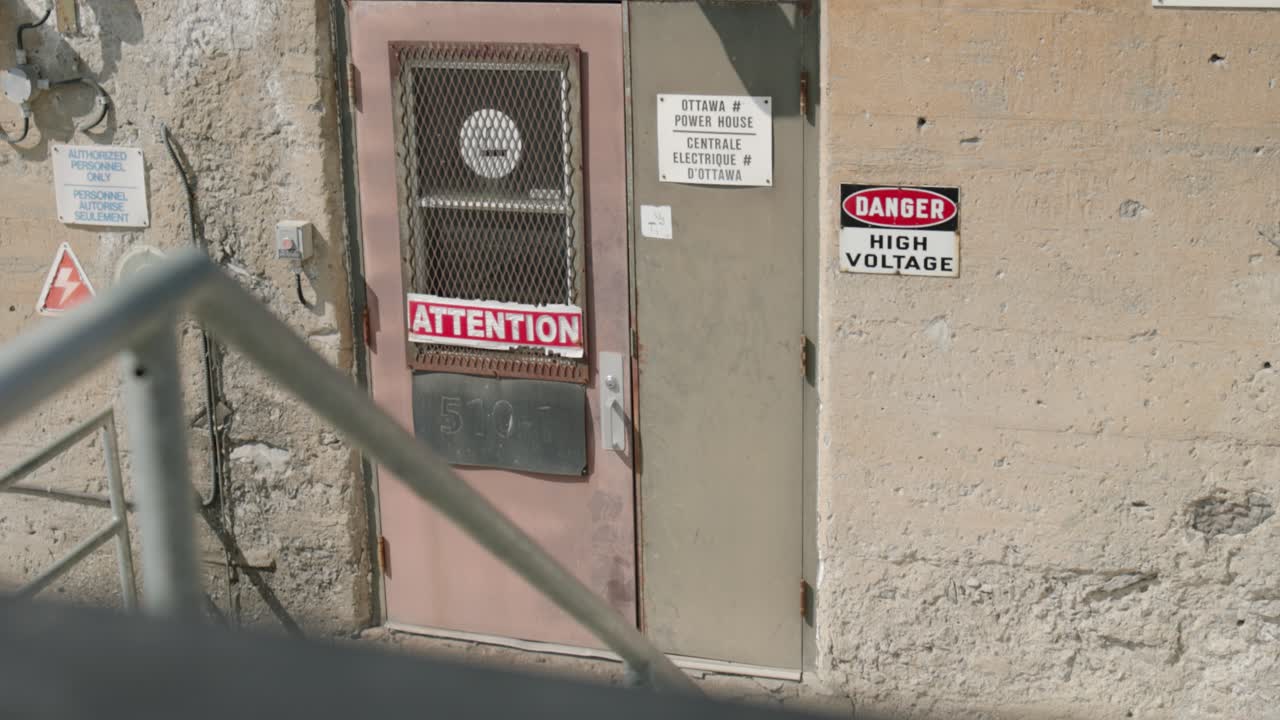 entorno de estilo industrial del exterior de una antigua instalación hidroeléctrica en la isla chaudière con señales de peligro de alto voltaje, puertas, barandillas y una escalera exterior