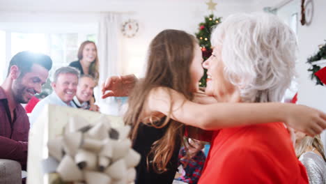 Nieta-Dándole-Un-Regalo-A-La-Abuela-Mientras-Una-Familia-De-Tres-Generaciones-Se-Sienta-En-El-Salón-Celebrando-Juntos-El-Día-De-Navidad-En-Casa