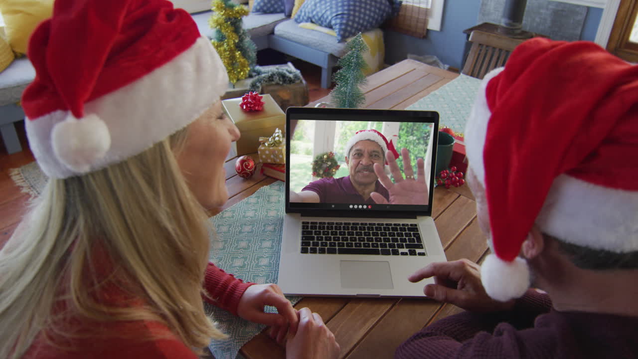 pareja caucásica sonriente con sombreros de santa usando una computadora portátil para una videollamada de navidad con un hombre en la pantalla