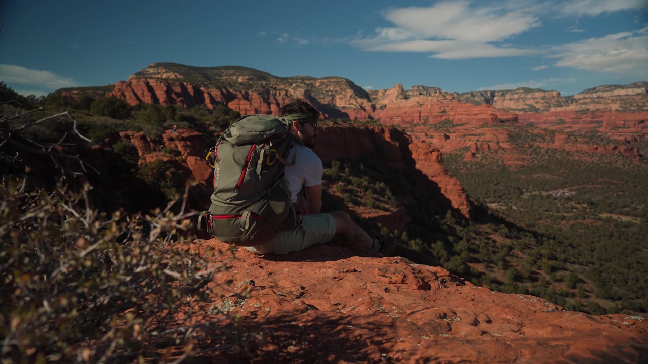 Wide shot of a male hiker walking to the edge of a red rock cliff in Sedona Arizona to sit and meditate