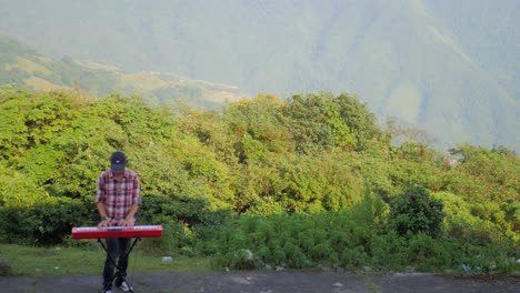 a stationary footage of a man, playing his electronic musical keyboard with a wide angle view of the mountain in the background. he also uses a pedal connected to his keyboard to change the tunes.