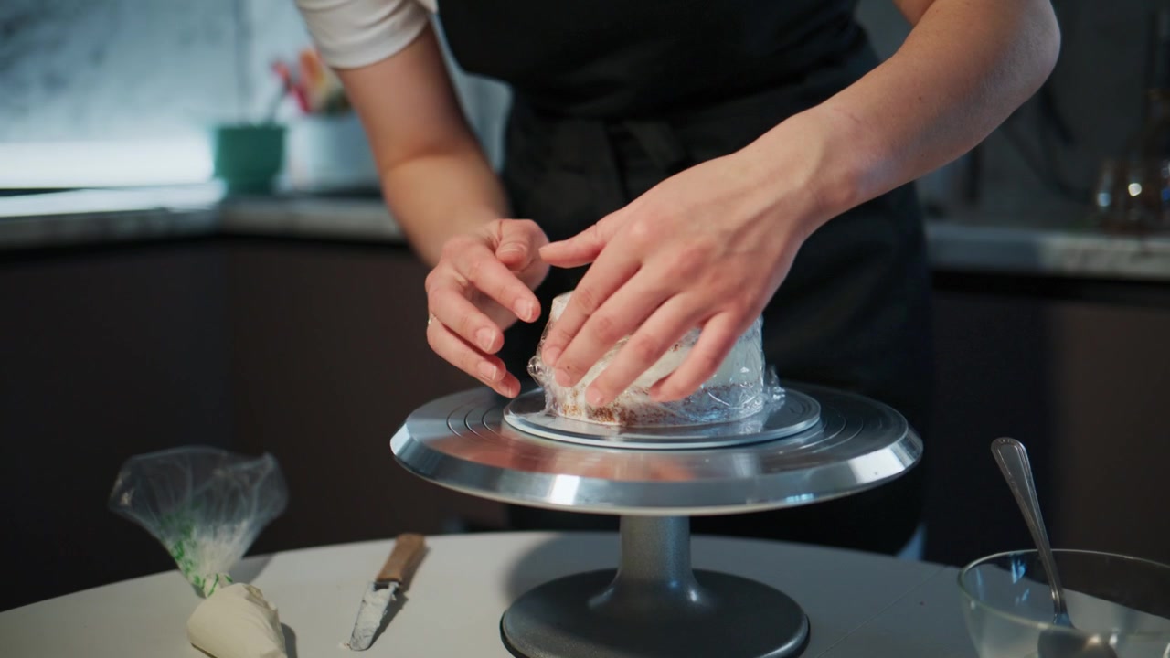 Focused young woman prepares a cake in a modern kitchen. She wears a black apron and white shirt. Her expression shows concentration and care for detail. The scene highlights baking, culinary arts
