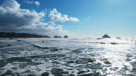 Impresionante-Toma-Aérea-De-Drones-De-4k-Deslizándose-Sobre-Las-Olas-Azules-Del-Océano-En-Bandon,-Oregon-Con-Nubes-Azules-Vibrantes