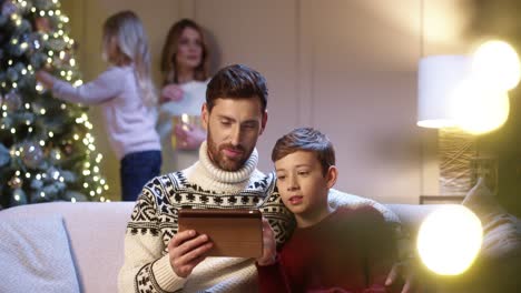 primer plano retrato de padre feliz con hijo adolescente sentado en la habitación tocando en la tableta eligiendo regalo de navidad en internet mientras mamá y linda hija decoran el concepto de vacaciones de año nuevo