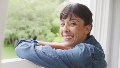 Portrait-of-happy-caucasian-woman-looking-through-window-and-wearing-jacket-in-living-room