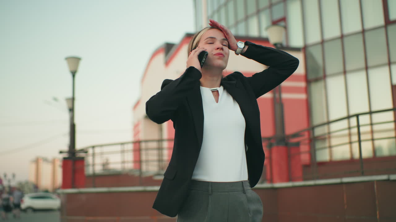 Frustrated white lady in professional outfit talking on phone in urban environment, looking tired with arm extended, conveying stress, emotional tension, and exhaustion