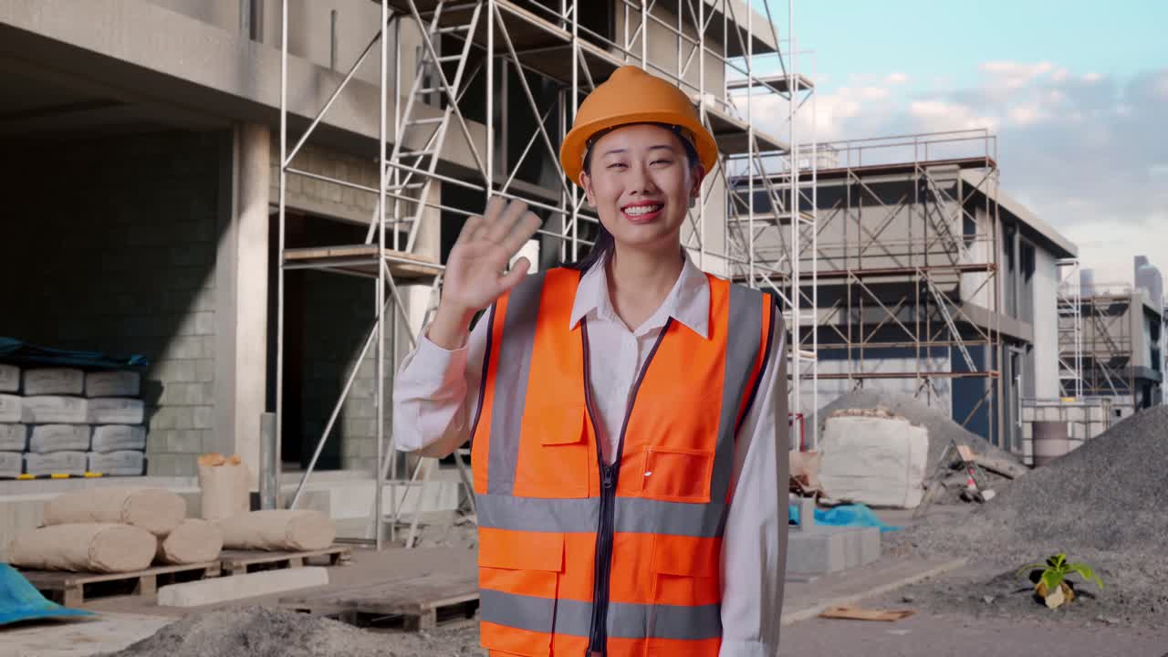 Asian Female Engineer With Safety Helmet Smiling To Camera And Waving Hand Saying Bye While Standing At Construction Site in Progress to New House