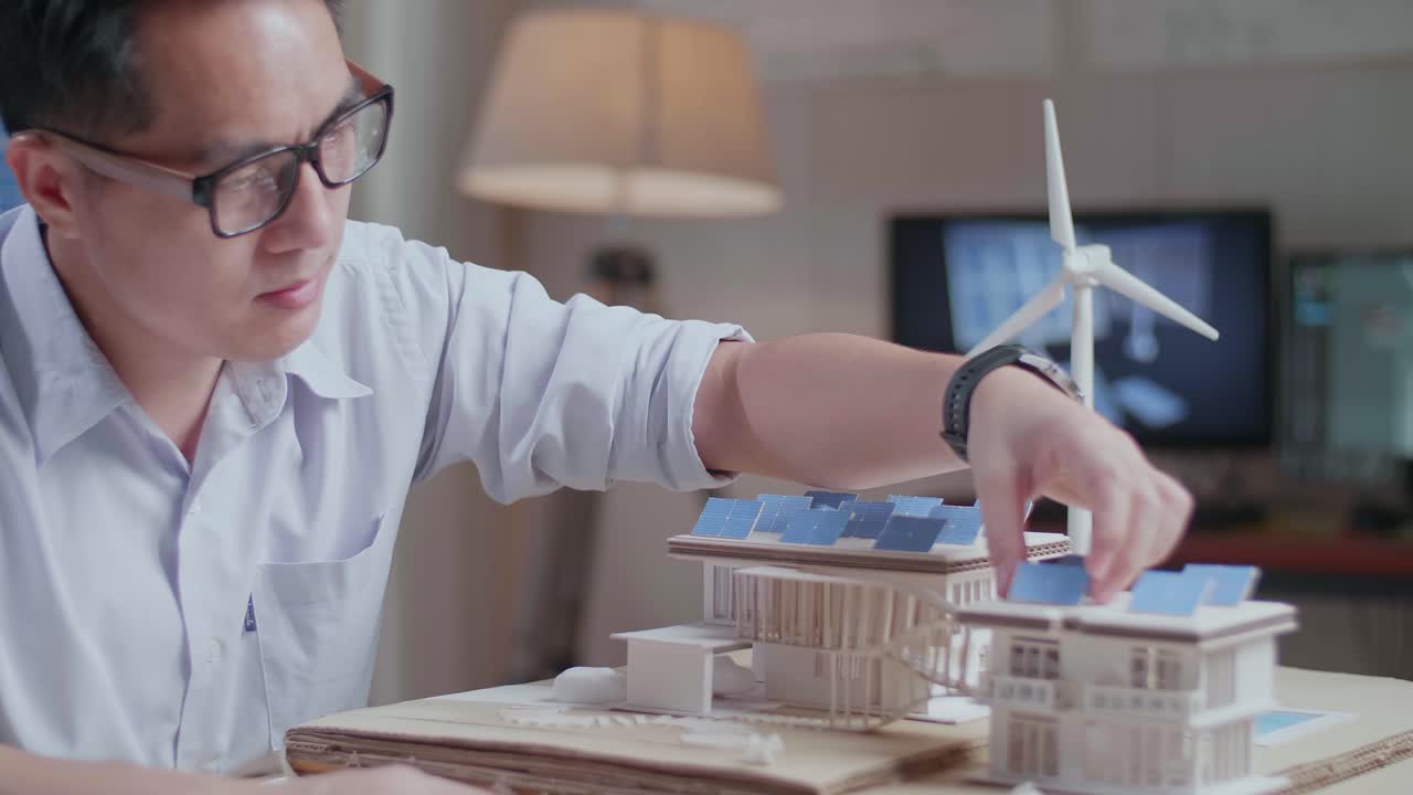 태양광 패널을 가진 작은 집의 모델을 건설하는 동안 아시아 남자가 긍정적으로 머리를 흔들고 있습니다.