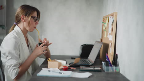 female nurse wearing glasses seated at desk drinking dark juice with straw during break, next to laptop and lunch box in bright modern office with corkboard full of notes and medical documents