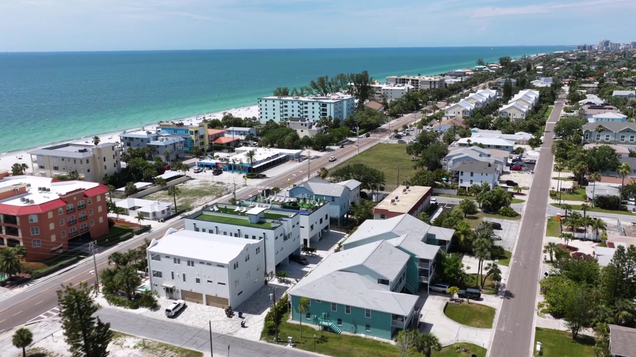 File di case fronte mare costeggiano la strada di Indian Rocks Beach, acque turchesi del Golfo incontrano sabbia bianca mentre strade fiancheggiate da palme si estendono verso lontani grattacieli costieri. Ripresa aerea con drone