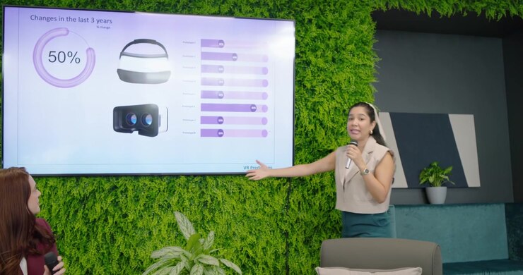 Two adult women holding mics in set, screen changing, right host standing and explaining VR 50%
