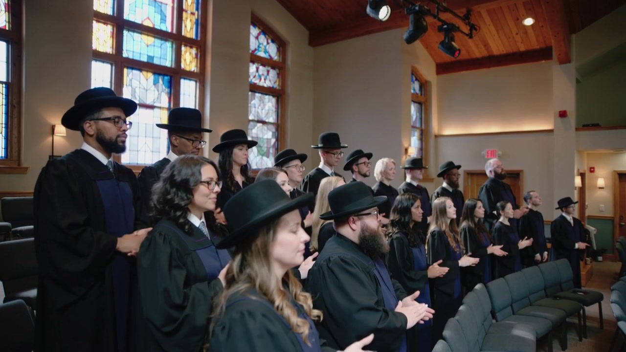 Na teken, zingend koor treedt op en klapt op verhoogde zitplaatsen in kapel, in gewaden en hoeden