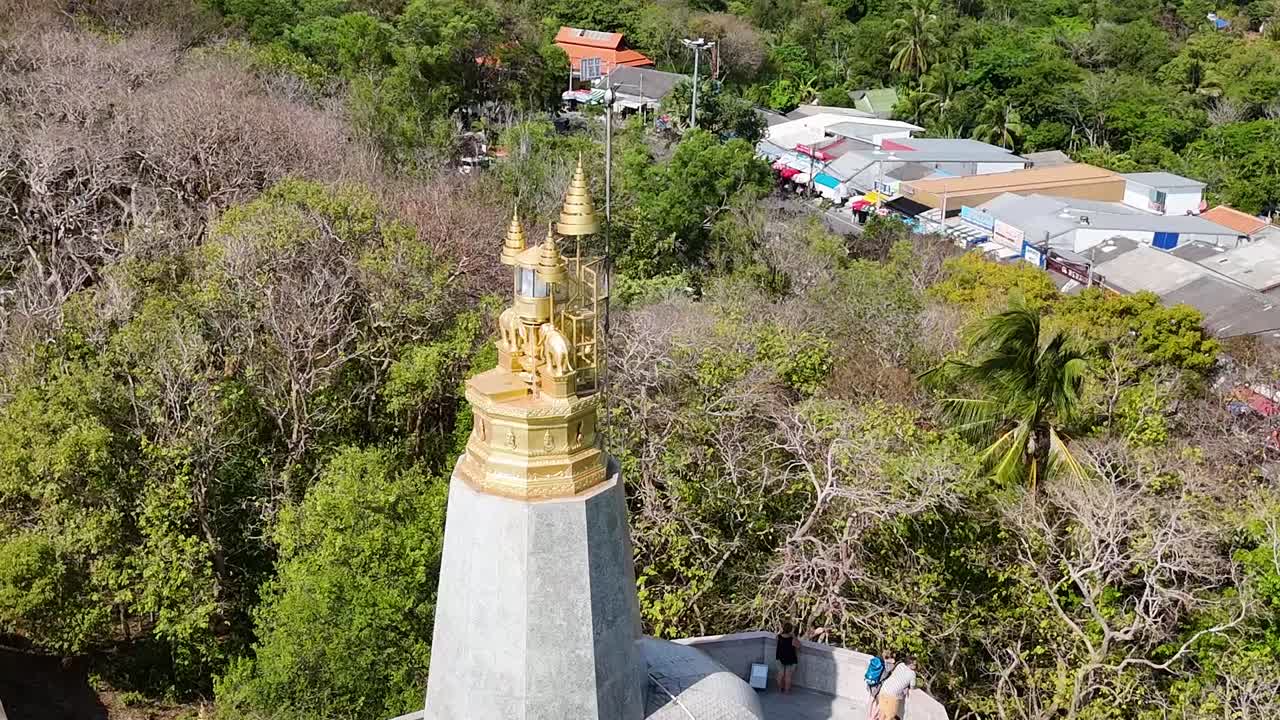 Aerial view of a golden spire surrounded by dense trees and distant hills under a clear sky.