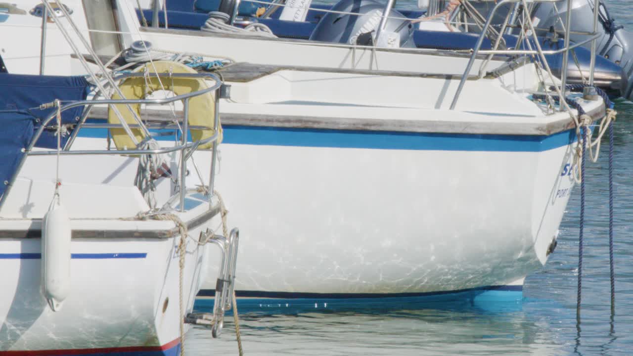 White sailboats remain stationary at a marina dock in Elie, Scotland, under bright daylight. Camera holds a steady, close-up view with gentle water reflections