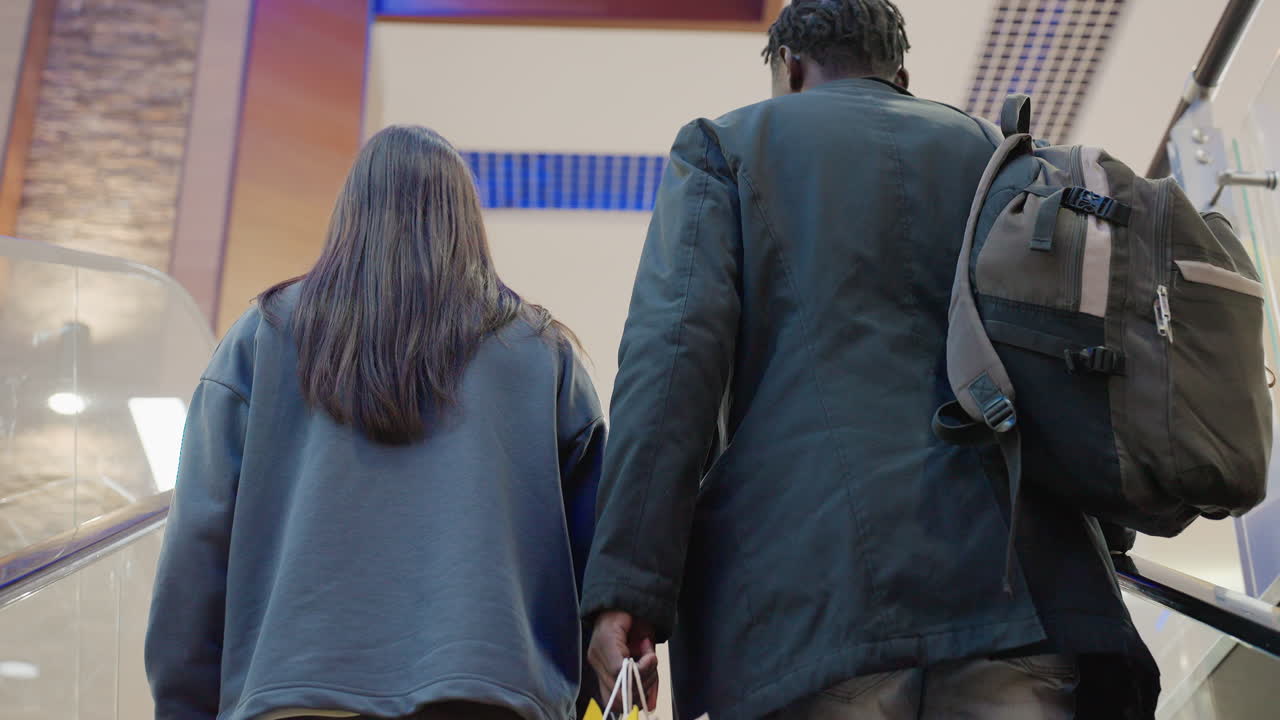 Back view of young man with backpack and woman with long hair holding shopping bag while ascending escalator inside modern mall, capturing quiet moment of shopping journey