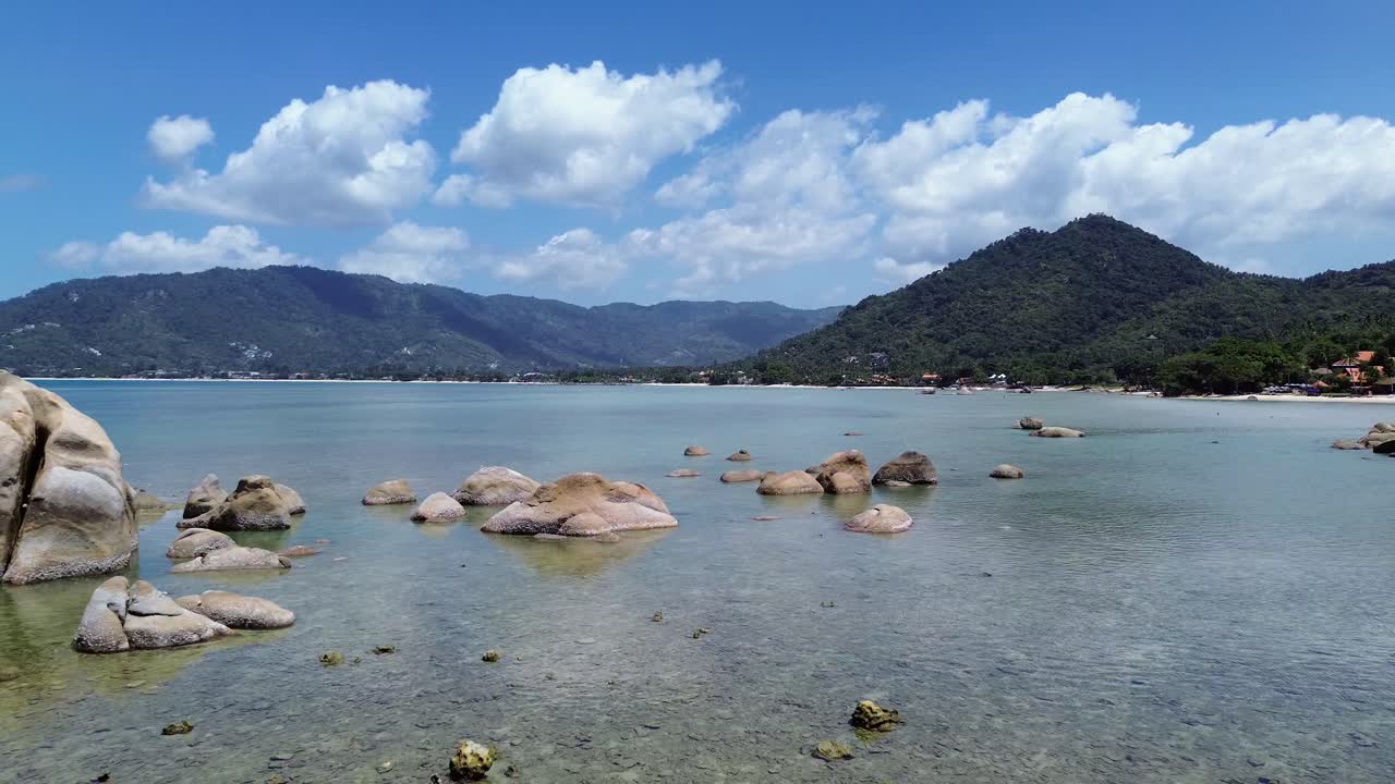 Aerial drone footage of Lamai Beach, Koh Samui, Thailand, showcasing rocky coastline, turquoise ocean water, lush green mountains, and scenic tropical island horizon under a clear sky