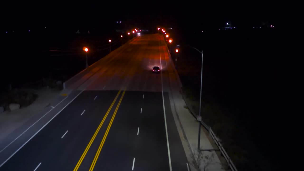 Aerial drone footage over a highway or a motorway on which a small gray or grey car vehicle is driving further during night time. Road has yellow and white marking on it and red, yellow street lamps.