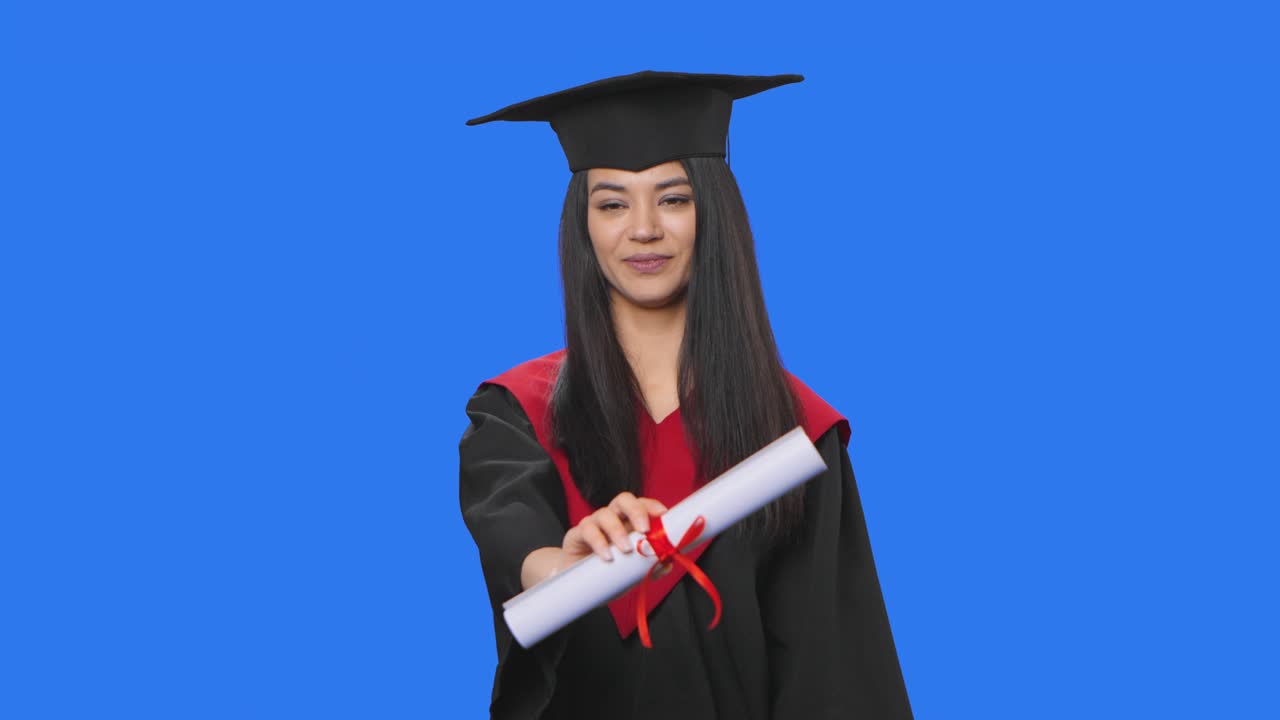 retrato de una estudiante en traje de graduación de gorra y túnica, sosteniendo un diploma y mostrando los pulgares hacia arriba. mujer joven posando en el estudio con fondo de pantalla azul. de cerca. cámara lenta lista 59.94fps