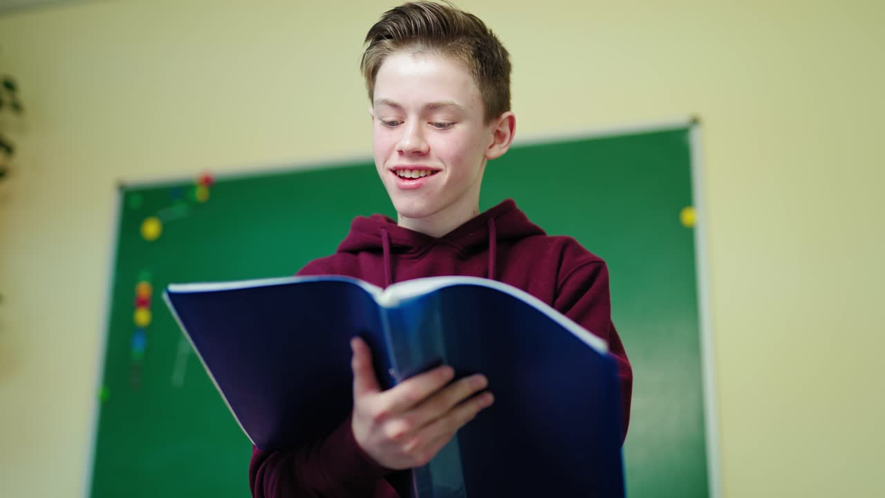 Smiling schoolboy with a folder. Teenage boy standing in the classroom and looking into the school register. Portrait of a happy teenager in the school.