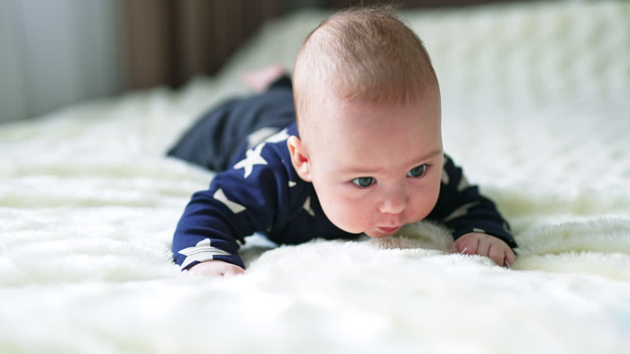 Nice cute little boy struggling to keep his head up. Kid in dark clothes left on a big white bed. Close up.