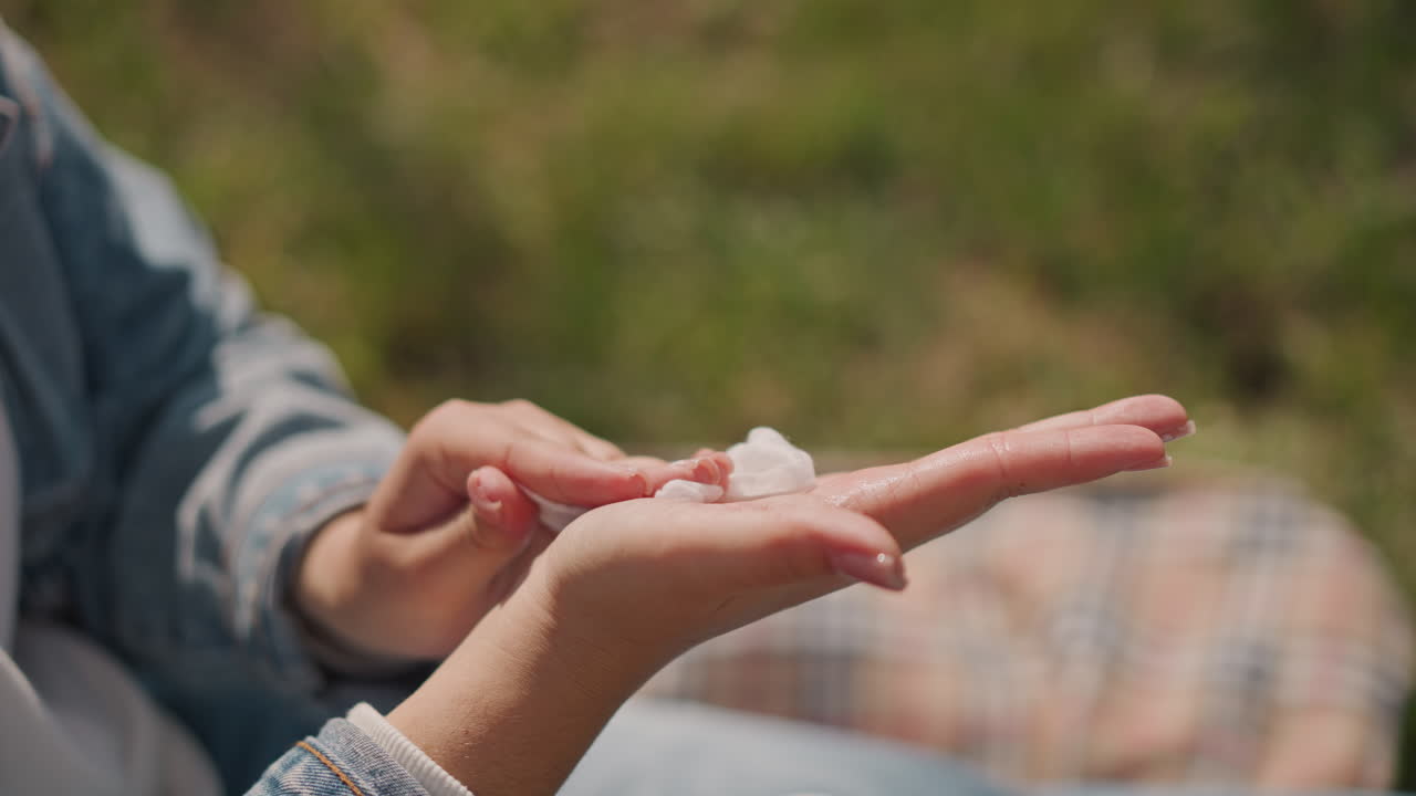 Closeup hands applying sunscreen cream outdoors picnic blanket, woman gently rubs lotion into palms and fingers, denim sleeve, soft sunlight, grassy meadow, relaxed summer selfcare moment