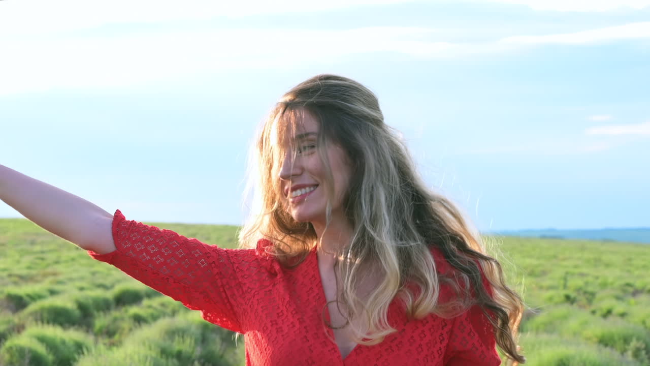 A woman in a red dress walks through a lush green field with the wind in her hair