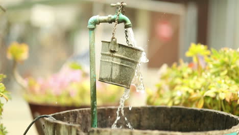 water from a garden spout pours and spills over a hanging bucket into a larger barrel