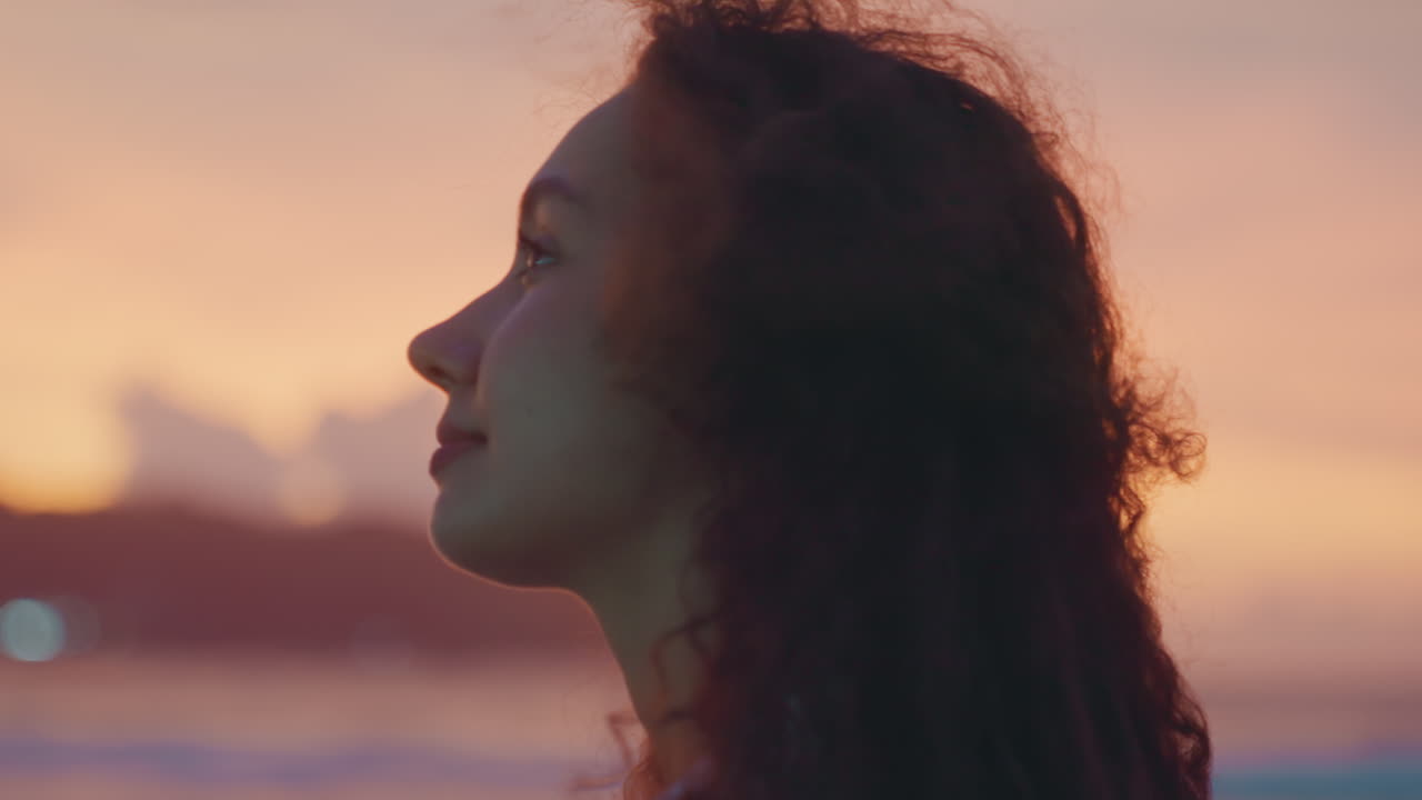 Mulher de cabelo cacheado com um sorriso gentil contemplando o oceano. Cenário de pôr do sol com mulher na costa arenosa com ondas. Mulher mestiça com cabelo cacheado sorri suavemente contra a silhueta da ilha ao crepúsculo