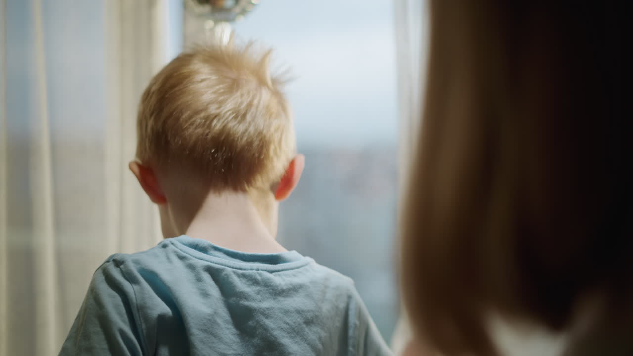 Smiling young boy in blue shirt looking sideways with joyful expression in warm sunlight indoors, face glowing with happiness while interacting with parent, capturing pure childhood innocence moment