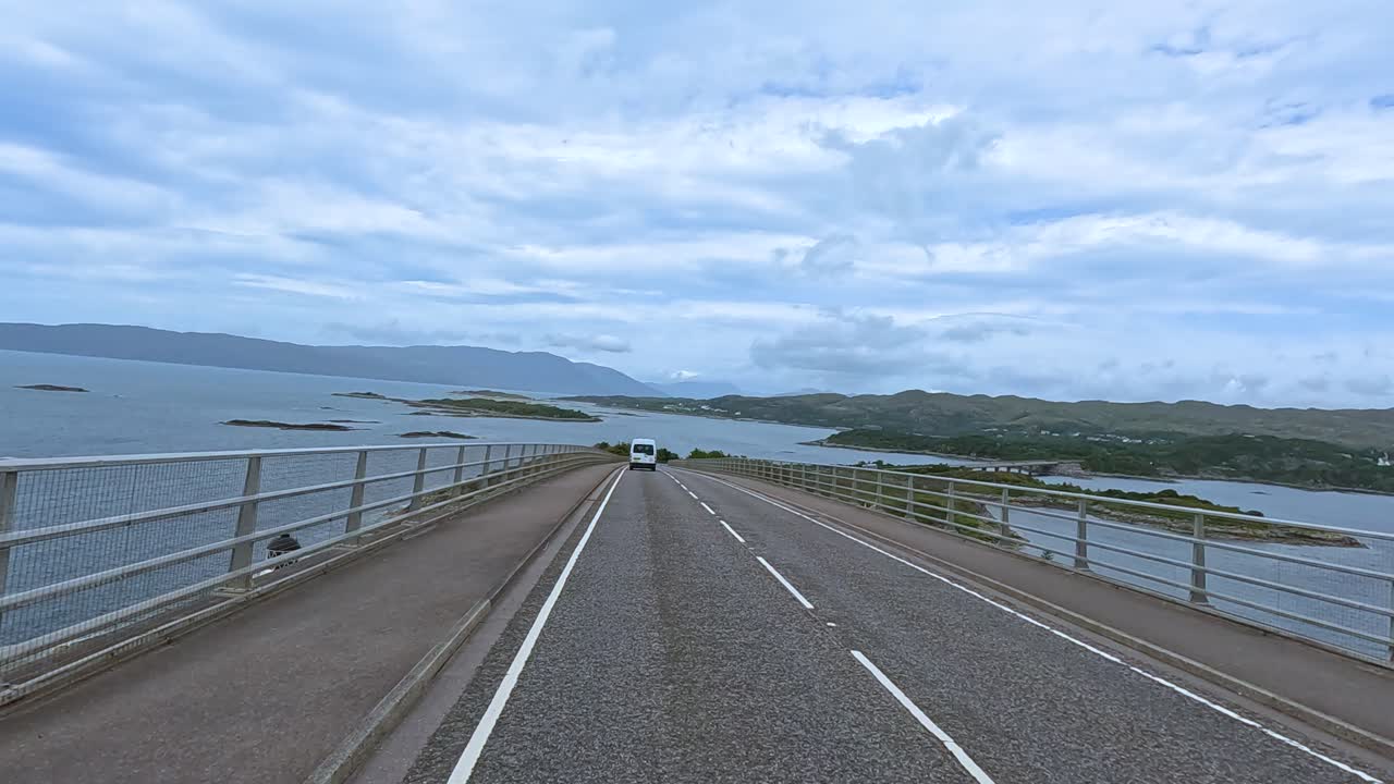 Forward-facing dashcam view of a vehicle crossing a bridge above water, with overcast daylight, distant hills, and steady camera movement