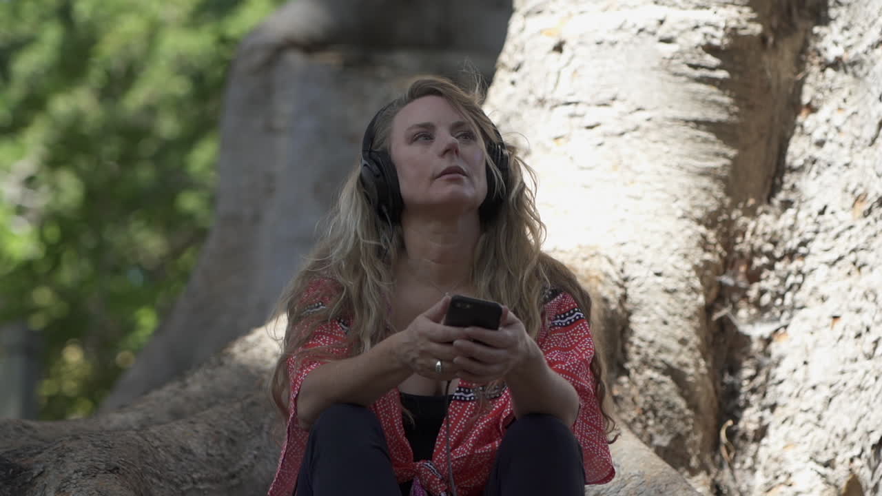 Attractive blonde woman sitting under tree in park while listening to music in headphones