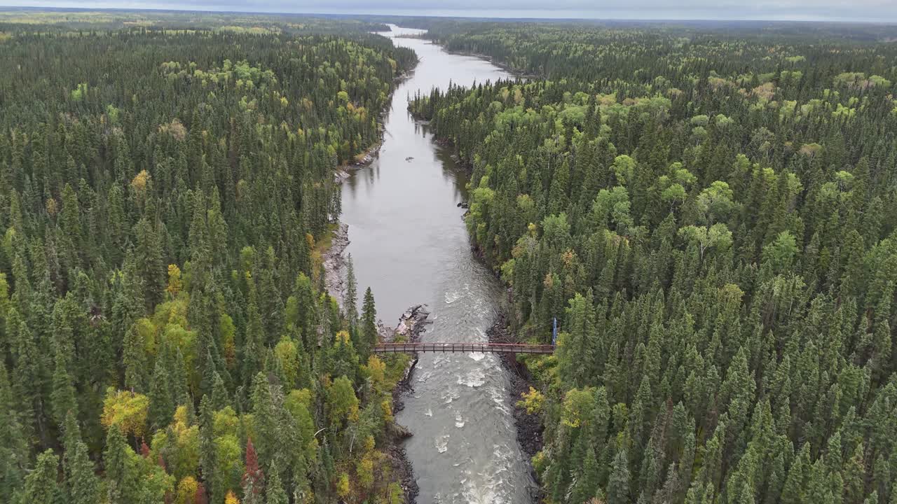Ilmadrone lentää korkealla Grass-joen yläpuolella Pohjois-Manitobassa, Kanadassa, seuraten sitä alavirtaan kohti riippusiltaa. Sillan alla näkyy koskia ja joen mutkia syksyisen lehtien väriloiston ympäröimänä.