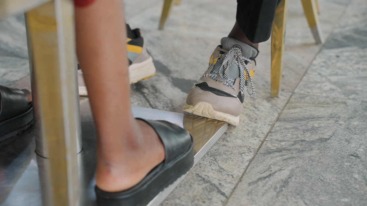 Gioco di piedi giocoso sotto il tavolo del caffè, coppia che si scambia un tocco segreto con i piedi durante un appuntamento rilassato all'aperto, primo piano di piedi e scarpe sotto sedie di metallo su pavimentazione piastrellata, sneakers casual e sandali in pelle