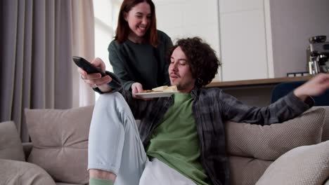 happy brunette guy with curly hair in a checkered shirt watches tv and uses the remote control while a brunette girl brings him snacks for the movie and communicates with the guy hugging him at home