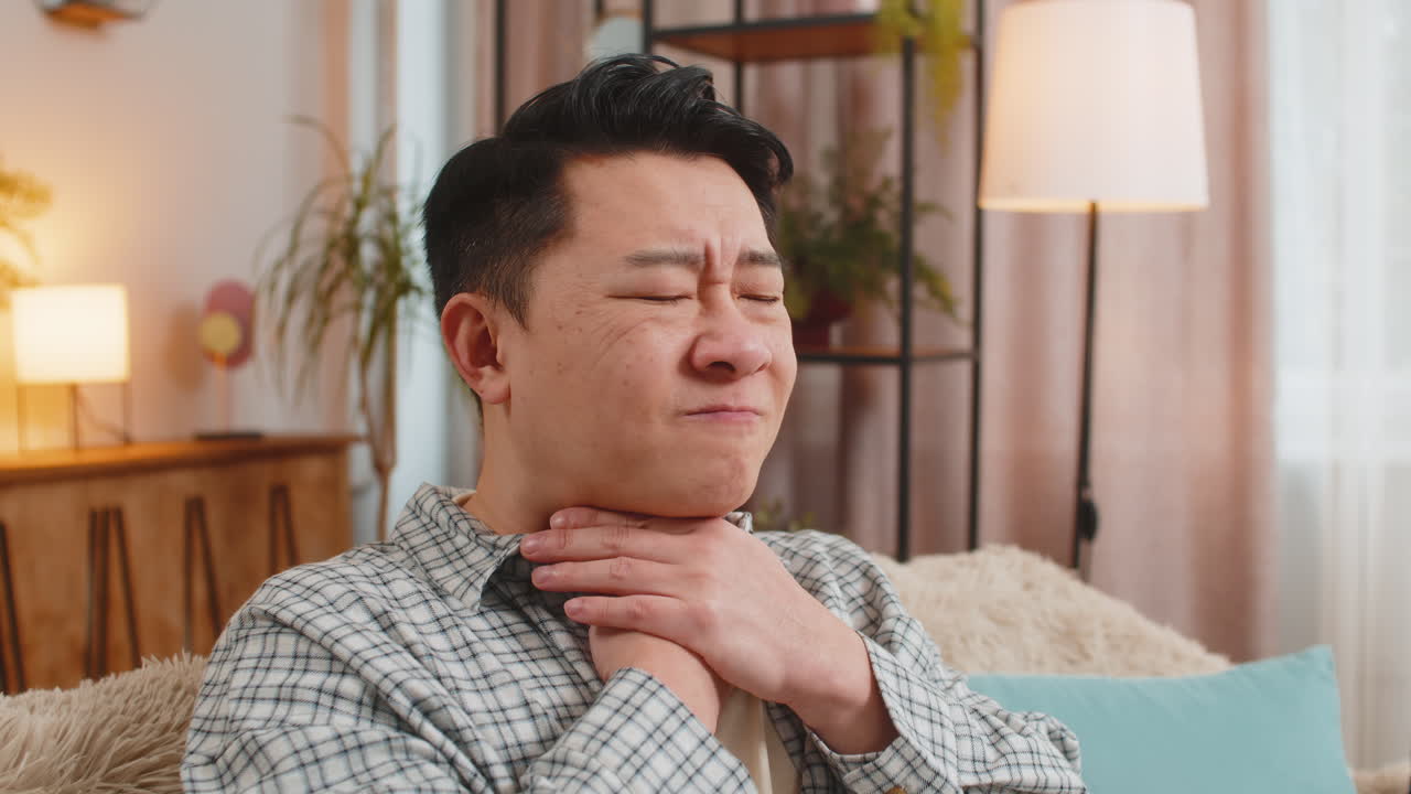 Upset chinese young man holds hands over sore throat with painful neck and frowning on sofa at home