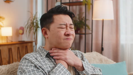 upset chinese young man holds hands over sore throat with painful neck and frowning on sofa at home