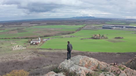 an orbiting aerial view of a man walking to the edge of a mountain top, looking out at the vast green field, with mountains, trees and sky in the background