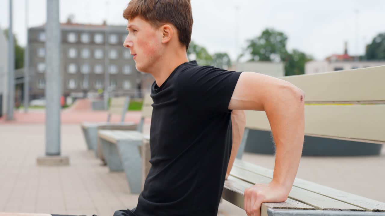 Young man performing tricep dips on a city park bench, staying active and building strength with calisthenics during an outdoor urban workout session, slow motion, static camera shot