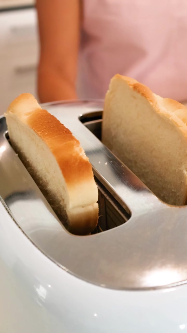 Slices of bread rise from a toaster in a bright kitchen. A person stands nearby, creating a warm, domestic atmosphere