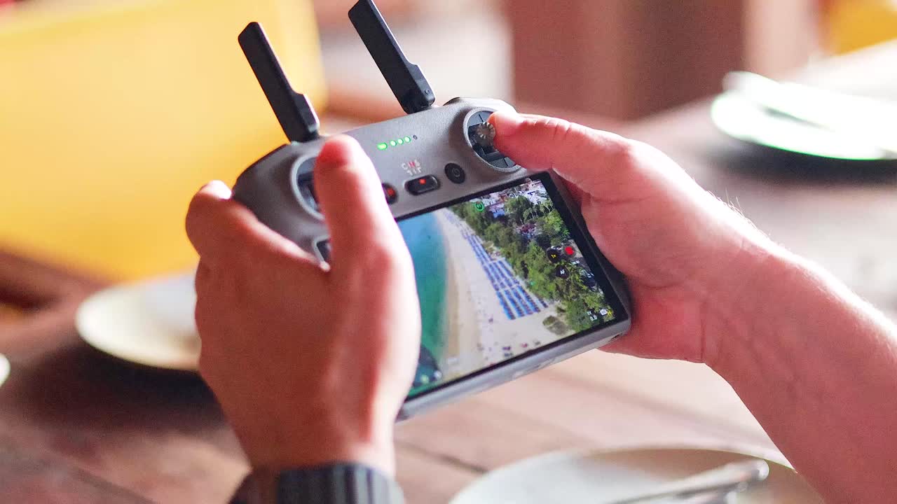 Hands operate a drone controller displaying aerial views of Phuket, Thailand. Bright indoor lighting highlights the device and screen