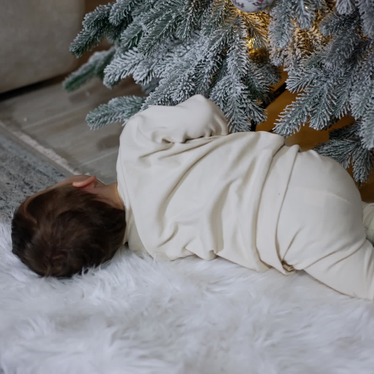 Beautiful toddler lies under Christmas tree waving his little hands. Baby rolls on the soft fluffy plaid trying to stand up