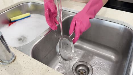 person wearing pink rubber gloves rinses kitchen utensils under running water in a modern kitchen sink, with bright lighting and steady camera angle