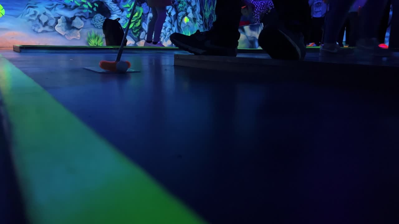 Mini golf club closes a white ball over a black mini golf course in a dark hall