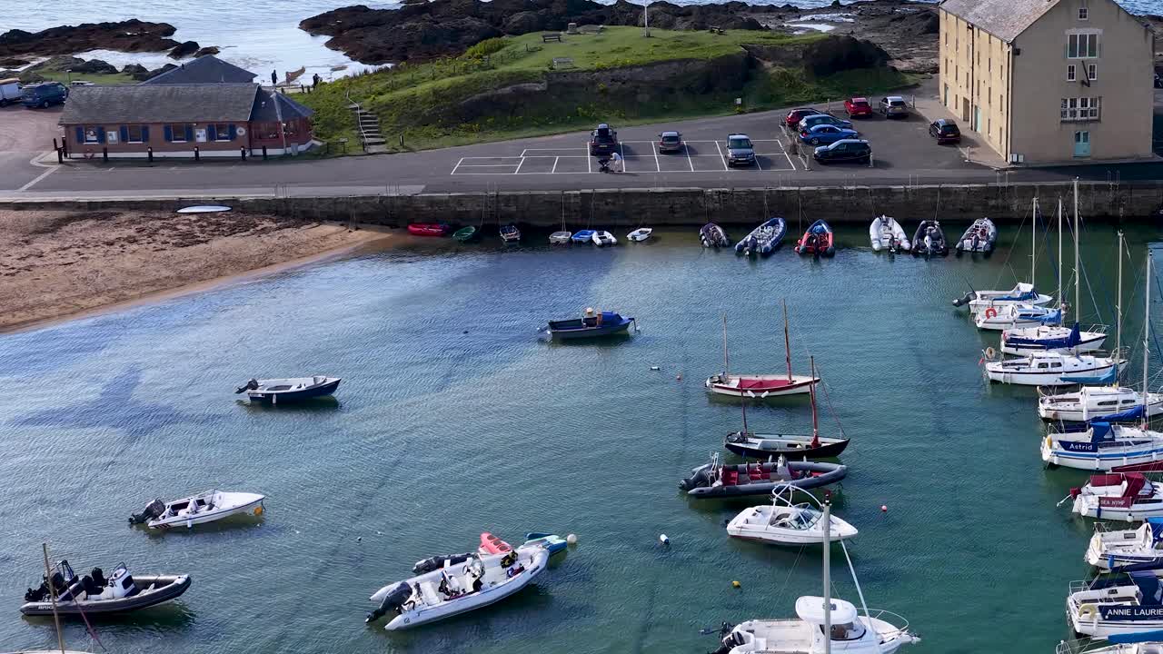 Drone glides over marina with moored boats, clear water, seaside buildings, and summer daylight