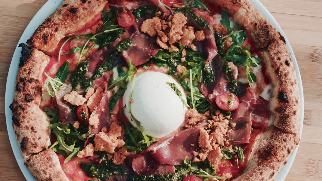 A beautifully shot overhead view of a gourmet pizza topped with burrata, prosciutto, cherry tomatoes, pesto, arugula, and crunchy crumbs