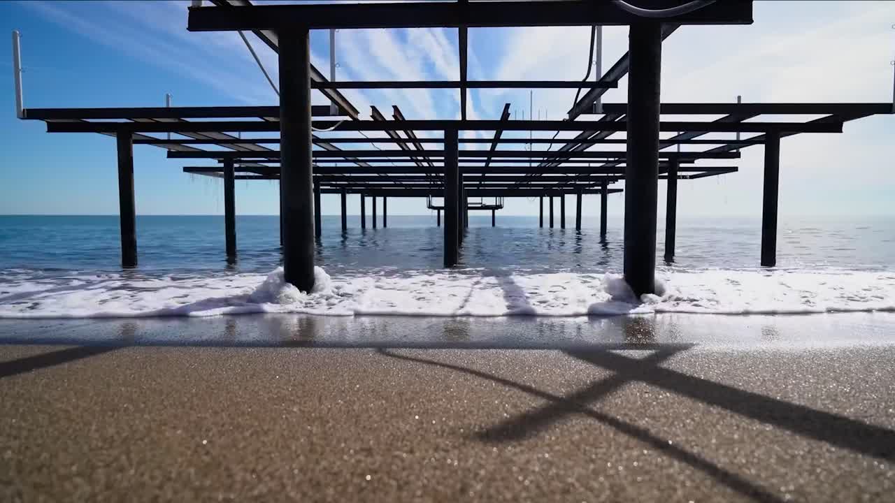 Big wooden or metallic harbor poles or masts in the beach while the sea and ocean water is hitting them in slow motion while the sun is shining and beaches sand is clearly visible in 4K resolution.