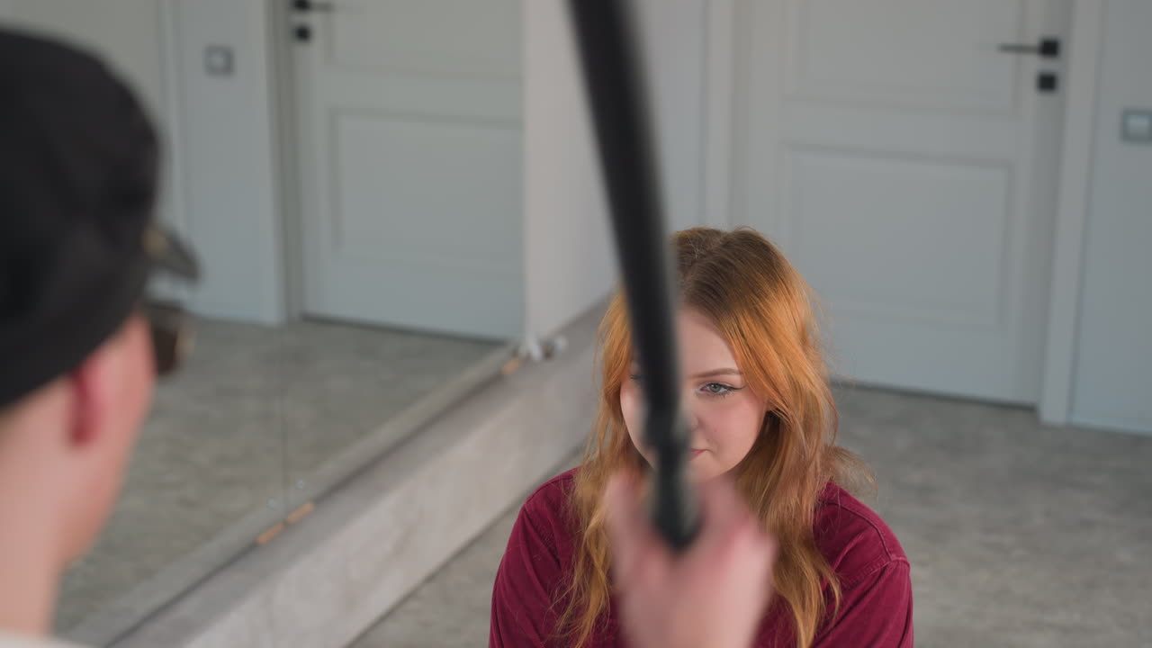 Rear view of man dancing playfully with stick in hand, approaching seated woman holding head while smiling. Lighthearted moment captured in indoor setting with warm lighting