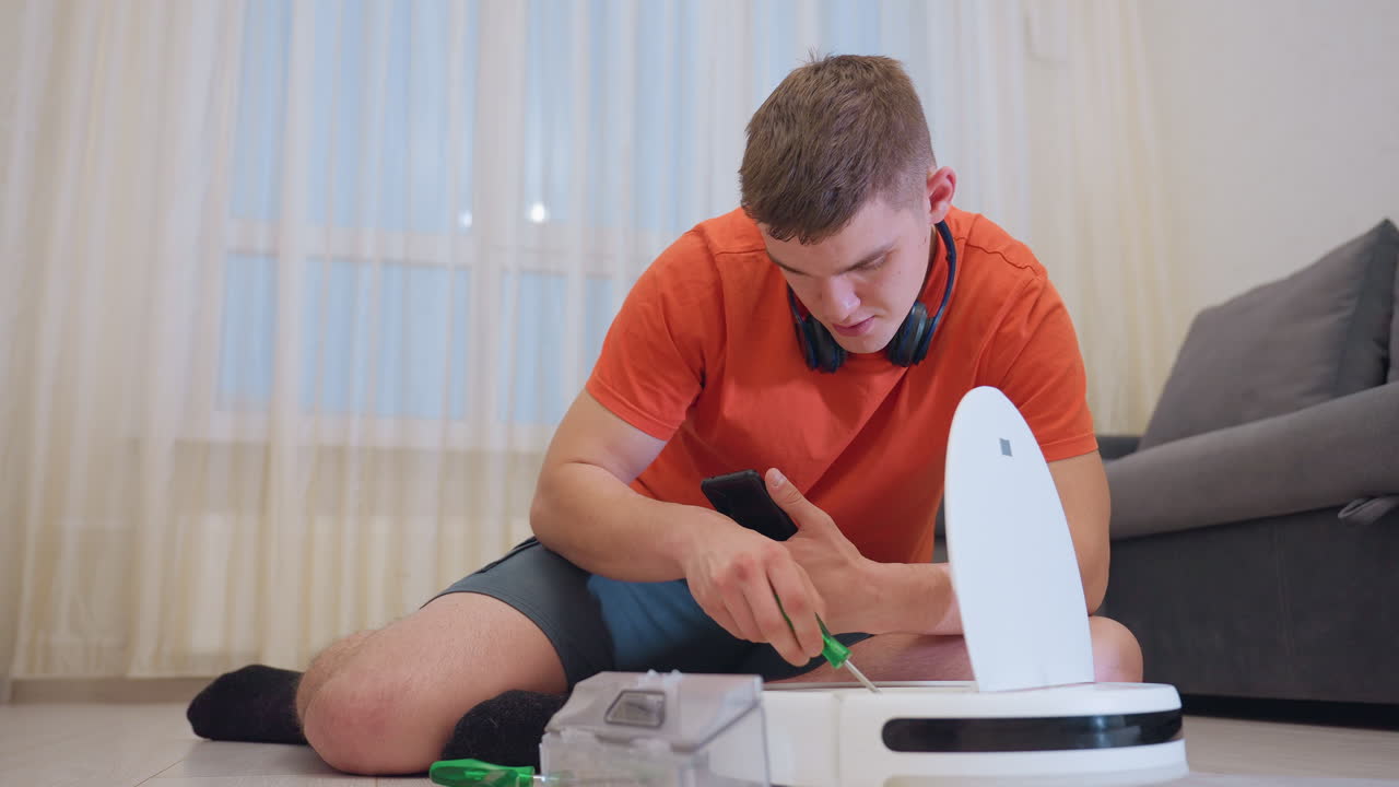 Man in orange shirt seated on floor beside sofa inspects robot vacuum cleaner using screw while holding phone, focusing on maintenance and repair of modern cleaning device with scattered tools