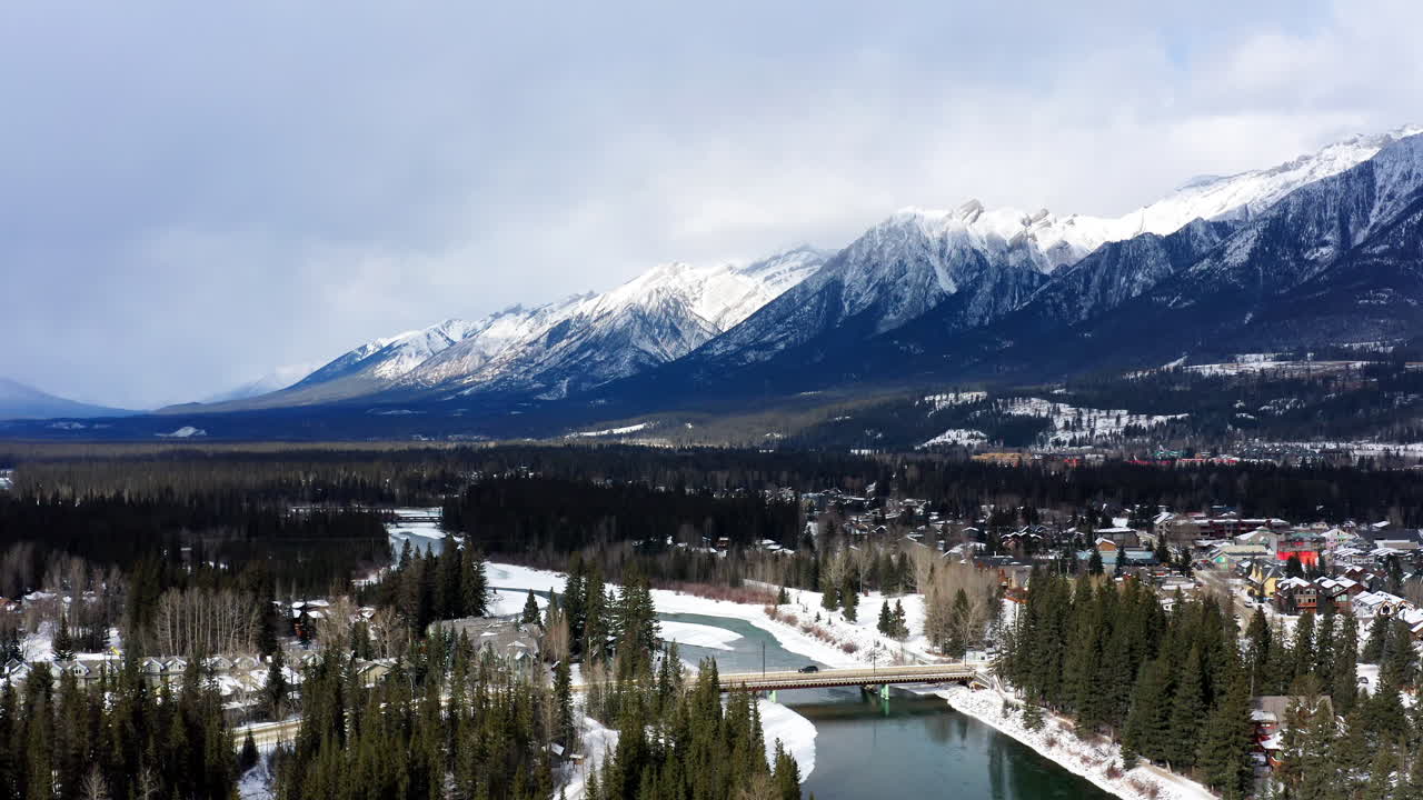 A wide drone fly-out captures the peaceful town of Canmore framed by snow-covered pines and the towering Banff mountain range.